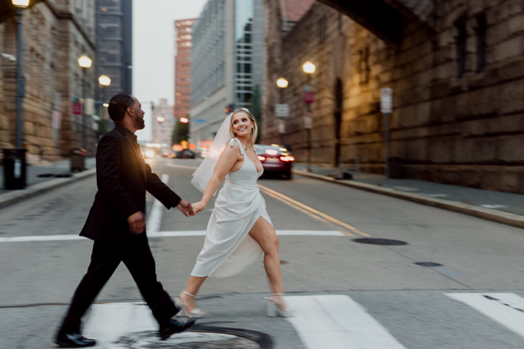 Couple eloping in Pittsburgh crosses a city street at dusk. The woman wears a white dress, and the man a black suit. Buildings and cars are visible in the background.